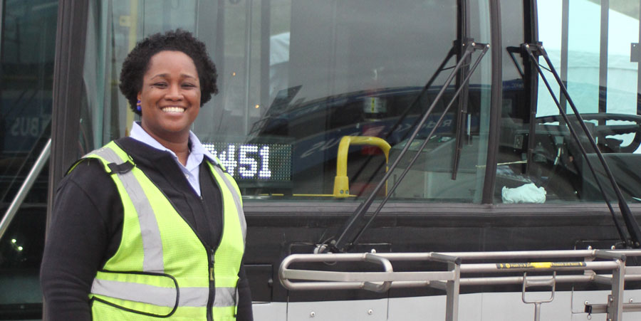 Bus driver in front of bus, smiling and wearing a safety vest.